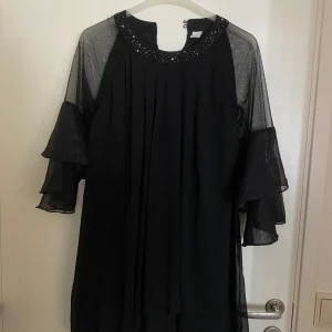 Svart klänning med paljettdetaljer - Elegant svart klänning med transparenta trekvartsärmar och paljetter runt halsringningen. Perfekt för festliga tillfällen. Klänningen har en lös passform och är tillverkad i ett lätt och luftigt material. storlek 170 men Passat XS-S det är små knappar i ryggen upp vid kragen! Så fin till fest! 