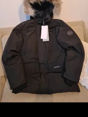 Svart Canada Goose dunjacka med päls - Svart dunjacka från Canada Goose med huva och snygg pälsdetalj. Jackan har flera fickor, patch på ärmen och dragkedja framtill. Perfekt för kalla vinterdagar och har en clean, stilren look som funkar till allt. Klassisk modell men liten i storleken. Det står Medium men jag skulle säga att den passar Small istället.