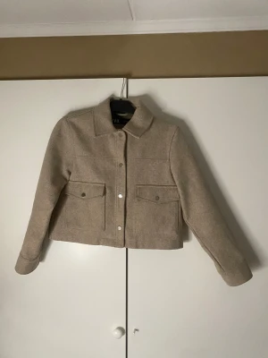 Beige kappa från Zara - Säljer en beige kappa från Zara i storlek S. Jackan har en croppad passform, klassisk krage, två stora fickor med lock och knappar framtill. Materialet är mjukt och känns ulligt, perfekt för lager på lager under höst och vår. Inga defekter!!