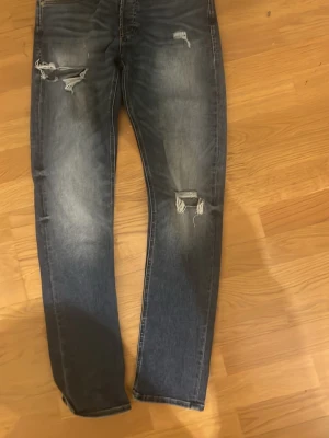 Slim fit jeans från Jack & Jones - Säljer ett par blå slim fit jeans från Jack & Jones, modell Glenn. Byxorna har slitna detaljer och hål på låret och knät.