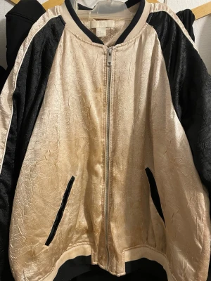 Beige och svart bomberjacka med struktur - Säljer en beige och svart bomberjacka från H&M med coolt strukturerat tyg och kontrastpaneler på ärmarna. Jackan har dragkedja framtill, svarta fickor och ribbade muddar. Perfekt för dig som vill sticka ut med en unik look.