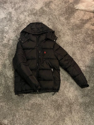 Svart pufferjacka från Polo Ralph Lauren - Svart pufferjacka från Polo Ralph Lauren med huva och dragkedja framtill. Jackan har två sidofickor med dragkedja och en röd Polo-logga på bröstet. Jackan är i storlek S och passar dig som är ca 178. Väldigt fint skick finns inga tecken på andvändning.