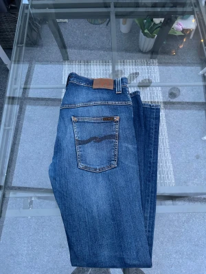 Nudie Jeans Tape Ted W32 L32 – Grey Worn Indigo - Säljer mina Nudie Jeans – Tape Ted i storlek W32 L32. Riktigt snygg tvätt i Grey Worn Indigo med naturligt slitage som ger en fin vintage-känsla. Jeansen är gjorda i 98% ekologisk bomull och 2% stretch, så de är både sköna och hållbara.  Modellen är slim utan att vara tight, och de ikoniska Nudie-bågsömmarna på fickorna gör dem lätta att känna igen. Inga hål eller skador – bara naturlig fading som ser grym ut.  Perfekta om du gillar premium denim, bra passform och tidlös stil.