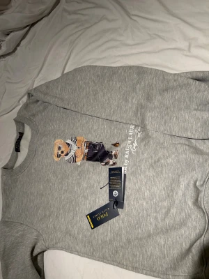 Grå sweatshirt Polo Ralph Lauren - Säljer en grå sweatshirt från Polo Ralph Lauren med Polo Bear-tryck på bröstet. Tröjan har rund hals, ribbade muddar och är långärmad. Materialet är mjuk bomull och polyesterblandning, perfekt för chill dagar. Klassisk och stilren design med coolt motiv. Ny pris 1899kr
