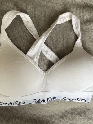 Vit linnetopp från Calvin Klein - Säljer en vit topp från Calvin Klein med breda korslagda axelband och elastisk logotypkant nedtill. Toppen är ärmlös och har en sportig design som passar perfekt till träning eller chill. Materialet är mjukt och stretchigt bomull för skön komfort. Passar 70-75 A/B