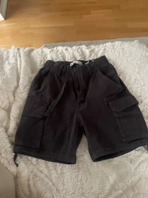 Mörkgrå cargo shorts från Bershka - Mörkgrå cargo shorts från Bershka med flera stora fickor på sidorna och baktill. Shorts med snörning i midjan och elastisk linning för skön passform. Tillverkade i bomull, perfekt för chill dagar. Snygg och enkel design som funkar till många stilar.