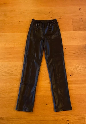 Svarta skinnbyxor från Nicki Studios - Säljer ett par svarta skinnbyxor från Nicki Studios med snygga snörningsdetaljer framtill. Byxorna har raka ben och hög midja, samt fickor bak. Materialet är mjukt och lite stretchiga. 