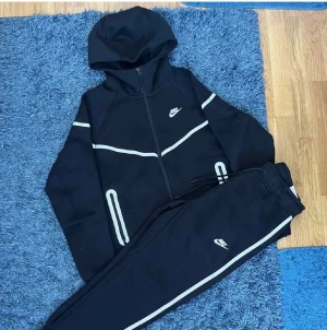 Svart Nike träningsset med huva - Säljer ett svart Nike träningsset med vit logga och vita detaljer. Setet består av en hoodie med dragkedja och matchande byxor med dragsko i midjan. Båda plaggen har vita linjer längs ärmar och ben för en sportig look. Perfekt för dig som gillar streetwear och sport.