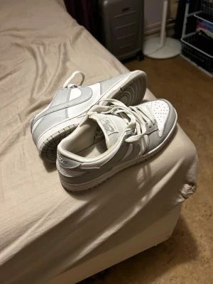Nike Dunk Low i vitt och grått - Nike Dunk Low sneakers i en clean vit och ljusgrå färg. Skorna har klassisk snörning, perforerad tå och rund tå. Ovandelen är i skinn och syntet, med den ikoniska swoosh-loggan på sidan. Perfekt för dig som gillar stilrena sneakers med låg profil.