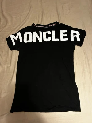Svart Moncler t-shirt med vit logga - Säljer en svart t-shirt från Moncler med stor vit logotyp över bröstet. T-shirten har klassisk rund hals och korta ärmar. Materialet är mjuk bomull som känns skönt mot huden. Perfekt för dig som gillar streetwear och vill sticka ut med ett tydligt märke.