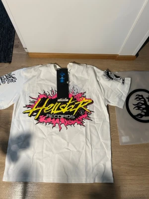Hellstar Records vit t-shirt med tryck - Säljer en vit Hellstar Records t-shirt med fet grafisk print i neonfärger och svart på framsidan, samt coolt tryck och text på ryggen. T-shirten har korta ärmar och är gjord i mjuk bomull. Extra detaljer på ärmarna med svart grafik. Perfekt för dig som gillar streetwear och statement-plagg.