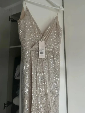 Glittrig långklänning till nyår - Säljer denna glittriga långklänning i ljus champagnefärg med smala axelband och omlott-design framtill. Klänningen har djup v-ringning, öppen rygg och en snygg slits som visar benet. Materialet är täckt av paljetter som ger en lyxig känsla. Jag har endast använt klänningen en gång vilket var på min bal, och lappen som sitter på insidan är till och med kvar! Den är helt felfri och som ny bortsett från på insidan längst ner på klänningen då jag kommit åt där med klackarna! 