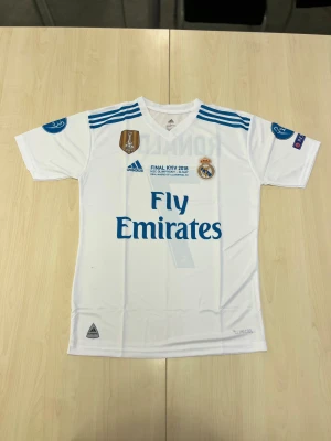 Real Madrid Football jerseys - Bilderna visar en vit Real Madrid fotbollströja med blå detaljer, Adidas logga och Ronaldo 7 på ryggen. 