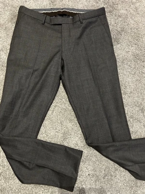 Mörkgrå kostymbyxor Oscar Jacobson - Snygga mörkgrå kostymbyxor från Oscar Jacobson i klassisk rak modell. Byxorna har bälteshällor och pressveck framtill, vilket ger en stilren och dressad look. Materialet är mjukt och känns lyxigt, perfekt för dig som gillar att klä dig stilrent.