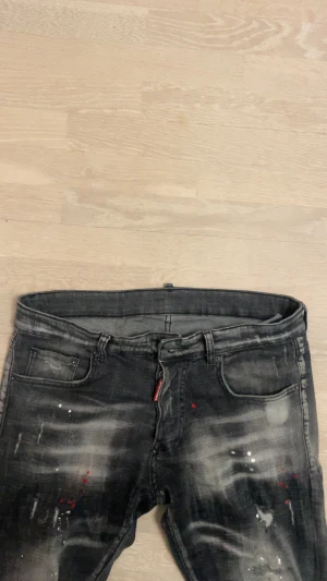 Svarta Dsquared2 jeans med färgstänk - Säljer ett par svarta jeans från Dsquared2 med coola slitningar och färgstänk i vitt och rött. Jeansen har fem fickor, smal passform och en snygg Dsquared2-patch bak.
