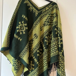 Grön och gul poncho med fransar - Poncho i mörkgrönt och gult med coolt geometriskt mönster och långa fransar längs kanterna. Perfekt att slänga över sig för en chill och unik look. Tjockt stickat material som håller dig varm under kyliga dagar.
