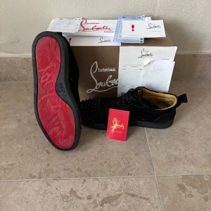 Christian Louboutin svarta sneakers med nitar - Svarta sneakers från Christian Louboutin i mocka med ikoniska röda sulor och svarta nitar på tån. Skorna har snörning, rund tå och lyxig läderinsida i beige. Kommer med originalkartong, dustbag och äkthetsbevis. Perfekta för dig som vill sticka ut med exklusiv stil till vintern. Stl 43 men passar 42-44 I perfekt skick utan några större skador