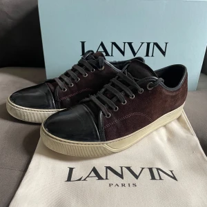 Lanvins  - Lanvin sneakers med mörkbrun mocka på sidorna och svart skinn på tå och plös. Skorna har rund tå, snörning och beige platt sula. Snygg kontrast mellan material och färger, perfekt för dig som gillar stilrena detaljer och exklusiv känsla. Storlek 7 us så alltså 40 i eu. Men dom upplevs större skulle säga att dom passar bäst för dom som har 43/44! Enda som behövs bytas är väl skosnörena som är lite slitna💯✅