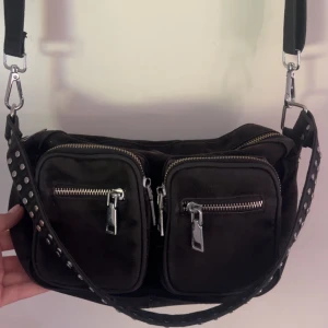 Svart axelväska från Noella - Cool svart axelväska från Noella i canvas med två stora framfickor och silvriga dragkedjor. Väskan har bred axelrem med nitar och flera fack inuti för extra förvaring. Perfekt för dig som vill ha en praktisk men snygg väska med lite edge. Använd men fortfarande i okej skick! Lite smutsigt inuti väskan och den är lite solblekt på utsidan som visas i bilderna💞nypris är runt 950kr