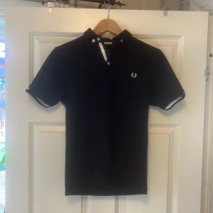 Svart piké från Fred Perry - Svart pike tröja från Fred Perry med vit broderad logga på bröstet. Skjortan har korta ärmar, vit knappslå och vita detaljer vid ärmslut. Tillverkad i mjuk bomull som känns skön mot huden. Perfekt för dig som gillar stilrena och klassiska plagg.