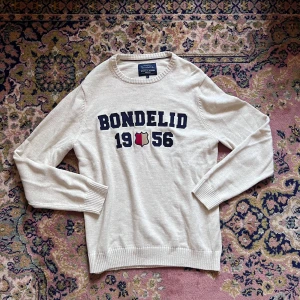 Beige stickad tröja från Bondelid - Snygg beige stickad tröja från Bondelid med mörkblå text och skölddetalj framtill. Tröjan har rund halsringning, ribbade muddar och lång ärm. Perfekt för dig som gillar klassisk och stilren look med en sportig touch.