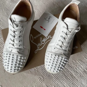 Vita sneakers från Christian Louboutin - Snygga vita sneakers från Christian Louboutin i skinn med ikoniska röda sulor och coola nitar på tån. Skorna har snörning och en klassisk låg siluett som sticker ut med sina detaljer. Perfekta för dig som vill ha något unikt och trendigt i garderoben.
