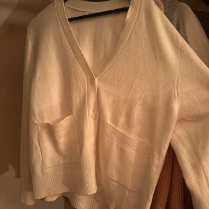 Vit kofta med fickor och knappar - Mysig vit kofta med v-ringning, knäppning framtill och två stora fickor. Tröjan har ribbade detaljer vid ärmslut och nederkant, vilket ger en stilren och enkel look. Perfekt att slänga på sig över en t-shirt eller linne.