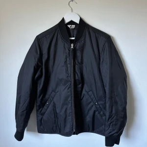 Svart bomberjacka från Arket - Svart bomberjacka från Arket med klassisk ribbad krage och muddar. Jackan är tillverkad i lätt nylon och har dragkedja framtill samt två snedställda fickor med tryckknapp. Fodrad med Thermore-isolering för extra värme. Perfekt för kyliga dagar. 3T2