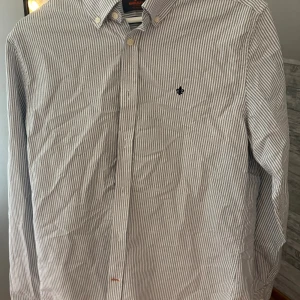 Randig skjorta från Morris  - Klassisk skjorta från Morris med smala vita och grå ränder, broderad svart logga på bröstet och knappar framtill. Skjortan har lång ärm och button down-krage, perfekt för en clean och stilren look. Aldrig använd helt ny