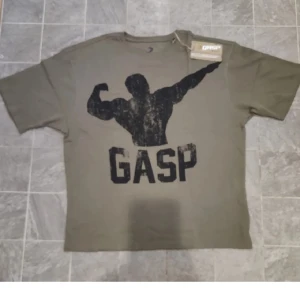 Olivgrön GASP t-shirt med tryck - Säljer en helt oanvänd olivgrön t-shirt från GASP med stort svart tryck av en bodybuilder och texten GASP på framsidan. Baksidan har motiverande text i svart. Klassisk passform, rund hals och korta ärmar. Tillverkad i bomull, perfekt för gymmet eller träning. 