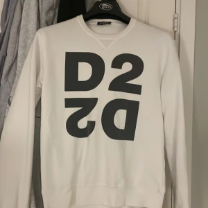 Vit sweatshirt från Dsquared2 - Säljer en vit sweatshirt från Dsquared2 med stor svart D2-logga på bröstet. Tröjan har rund halsringning och långa ärmar. Perfekt för dig som gillar stilrena och trendiga plagg med tydlig logga.