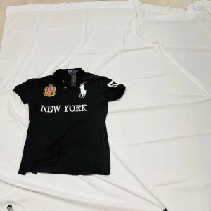 Svart Ralph Lauren t-shirt New York - Svart t-shirt från Ralph Lauren med broderad logga, New York-tryck och stort nummer 5 på ryggen. Klassisk pikékrage och korta ärmar. Materialet är bomull och detaljerna är broderade i vitt och guld.