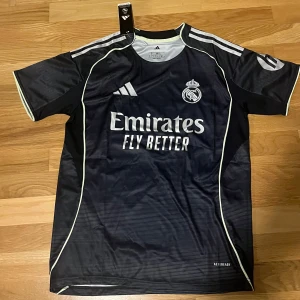 Real Madrid bortatröja 25/26 - Snygg Real Madrid bortatröja från Adidas i svart med tunna horisontella ränder och vita detaljer. Tröjan har klubbmärke, sponsortryck och HP-logga på ärmen. Tillverkad i lätt och ventilerande material, 