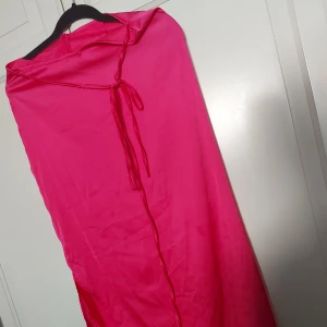 Lång rosa satinkjol med knytband - ⚠️ Skriv till mig innan köp⚠️Säljer en lång, glansig rosa kjol i satinliknande material. Kjolen har ett snyggt knytband framtill och en diskret dragkedja i sidan. Perfekt för dig som vill sticka ut med en färgstark och elegant look.