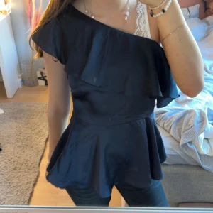 Mörkblå oneshoulder volangtopp i satin - Supersnygg svart topp i satin med oneshoulder och volangdetalj över axeln. Modellen har en markerad midja och utsvängd nederdel som ger en feminin siluett. Perfekt för dig som vill sticka ut med en trendig och elegant look. Accepterar dem flesta buden! Hör av er vid frågor!