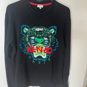 Svart Kenzo sweatshirt med tiger - Svart sweatshirt från Kenzo med ikonisk broderad tiger i grönt, blått och vitt framtill samt röd Kenzo-logga. Tröjan har rund hals, ribbade muddar och lång ärm. Perfekt statement-plagg för dig som gillar att sticka ut.