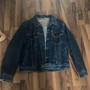 Levi's blå jeansjacka i storlek L - Klassisk blå jeansjacka från Levi's i storlek L. Jackan har snygga kontrastsömmar, bröstfickor med knappar och en tidlös design. Perfekt för dig som vill ha en ikonisk denimlook med cool vintagekänsla.