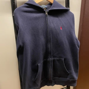 Mörkblå hoodie från Polo Ralph Lauren - Mörkblå hoodie från Polo Ralph Lauren i storlek 18-20 år. Tröjan har dragkedja framtill, huva och två stora fickor. Klassisk röd broderad logga på bröstet. Perfekt för dig som gillar en enkel och stilren look.