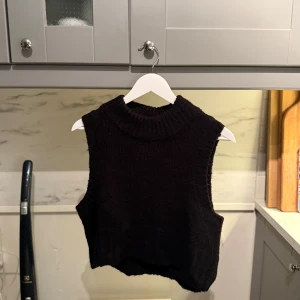 Svart stickad väst - Svart stickad väst med grov ribbstickad hög krage och ärmlösa kanter. Modellen är croppad och har en loose passform. Den är från VILA. 