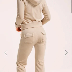 Beiga mjukisbyxor med fickor - Snygga beiga mjukisbyxor med två bakfickor med lock och guldiga knappar. Byxorna har en bekväm midja och är tillverkade i mjukt material som passar perfekt för en avslappnad stil. säljer både tröjan och byxorna 