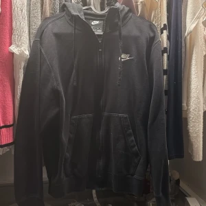 Svart Nike hoodie med dragkedja - Svart hoodie från Nike med broderad logga på bröstet, huva med snörning och praktiska fickor framtill. Tröjan har dragkedja hela vägen och är tillverkad i mjuk bomullsblandning. Perfekt för en avslappnad och sportig stil.
