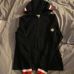 Svart Moncler hoodie med dragkedja - Svart hoodie från Moncler med dragkedja framtill och klassisk logga på bröstet. Huva och muddar har snygga ränder i rött, vitt och blått. Tillverkad i mjuk bomull, perfekt för en stilren och sportig look. Storleken är XL men sitter som en M
