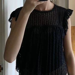 Svart spetsklänning från Nelly XS - Supersöt svart kortklänning från Nelly i spets med volangdetaljer på axlarna och knytning i nacken. Klänningen är ärmlös och har en transparent, mönstrad spets över hela plagget. Perfekt för dig som gillar romantiska och trendiga vibes.
