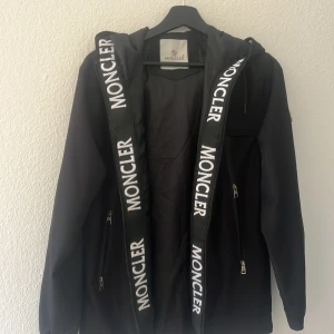 Svart vindjacka från Moncler med huva - Svart vindjacka från Moncler med huva och stora logoband längs dragkedjan. Jackan har dragkedjefickor, klassisk Moncler-patch på ärmen och coolt serietidningstryck på insidan. Perfekt för dig som vill ha en stilren och sportig look. Skannar du qr koden kommer du in på monclers hemsida.