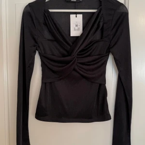 Svart omlottopp från Bik Bok - Säljer en svart omlottopp från Bik Bok med lång ärm och snygg draperad front. Toppen har en djup v-ringning och är figurnära i modellen. Materialet är tunt och stretchigt, perfekt för att skapa en trendig look.