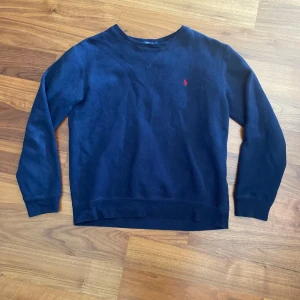 Marinblå tröja från Ralph Lauren - Säljer en klassisk marinblå tröja från Ralph Lauren med rund halsringning och diskret röd logga broderad på bröstet. Tröjan har långa ärmar och är tillverkad i mjuk bomull, perfekt för lager-på-lager-stil.