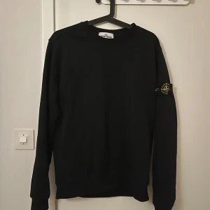 Svart sweatshirt från Stone Island - Svart sweatshirt från Stone Island med klassisk rund hals och ikonisk logotyp-patch på vänster ärm. Tröjan har en clean look och är tillverkad i mjuk bomull, perfekt för en stilren och avslappnad vibe.