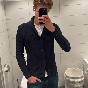 Mörkblå kabelstickad kofta John Henric - Säljer en mörkblå kabelstickad kofta från John Henric. Jag på bilden är 187 cm. Koftan har snygg knäppning med fyra knappar, ribbade muddar och klassisk krage. Perfekt för lager-på-lager och chill stil. Passar dig som gillar stilrena plagg. Tveka inte på att höra av dig vid prisförslag! Eller mer bilder