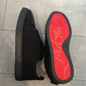 Christian Louboutin svarta sneakers med nitar - Säljer ett par svarta sneakers från Christian Louboutin med klassisk röd sula och coola svarta nitar på tån. Skorna har snörning och är gjorda i mocka med läderdetaljer. Perfekta för dig som vill sticka ut med en edgy och lyxig stil.