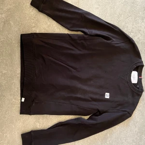 Svart sweatshirt från Les Deux - Svart sweatshirt från Les Deux med rund halsringning och ribbade muddar. Liten broderad patch med blått H på bröstet och diskret detalj i rött, vitt och blått vid halsen. Perfekt basic-tröja med clean look.